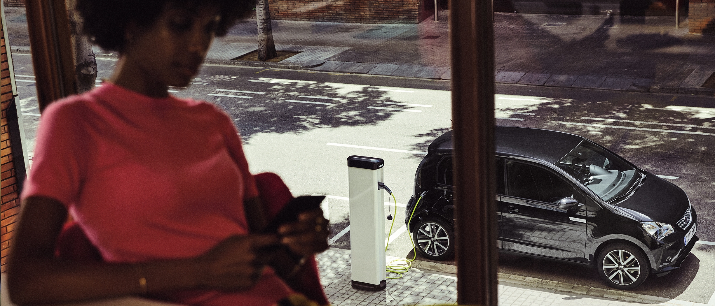 Op de voorgrond staat een vrouw met haar mobiel, achter haar staat een SEAT Mii Electric aan de lader