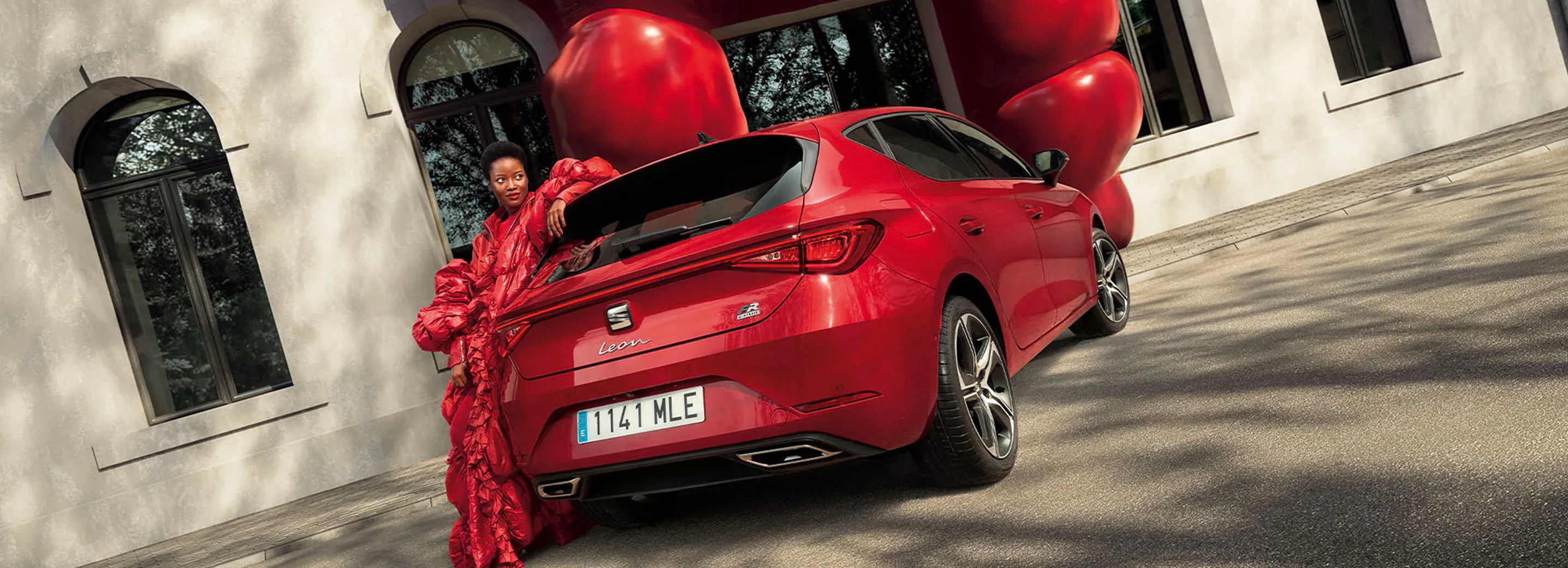 Vrouw in rood gewaad naast rode SEAT Leon